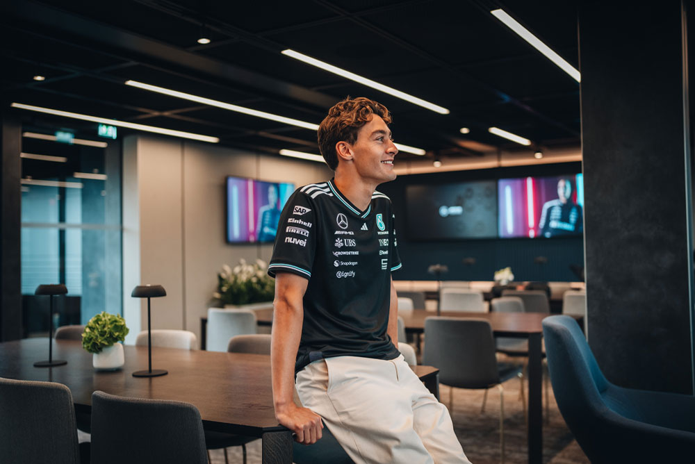 George Russell, fører for Mercedes-AMG PETRONAS Formula One Team, sitter avslappet på et bord i et moderne og stilfullt teamområde eller kontor. Han smiler og har på seg det offisielle teamskjortet med sponsorlogoer som Einhell, IWC og CrowdStrike synlige. Skjermer i bakgrunnen viser bildet hans.
