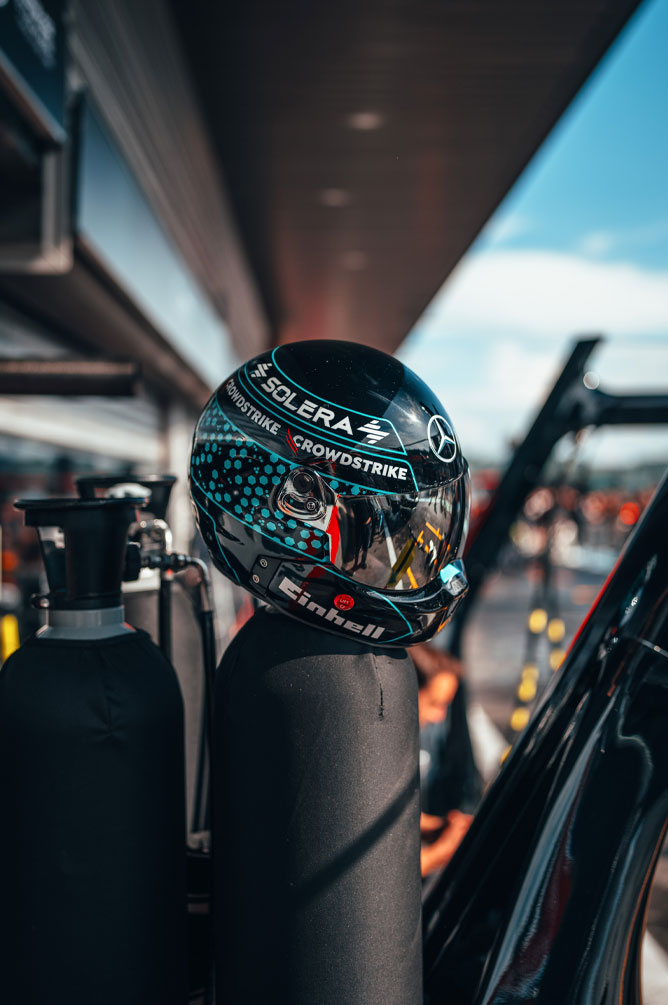 En hjelm fra Mercedes-AMG PETRONAS Formula One Team med tydelige sponsorlogoer, inkludert Einhell, CrowdStrike og Solera, er plassert på en gassflaske i pit lane. Refleksjonen i visiret viser omgivelsene i garasjen.