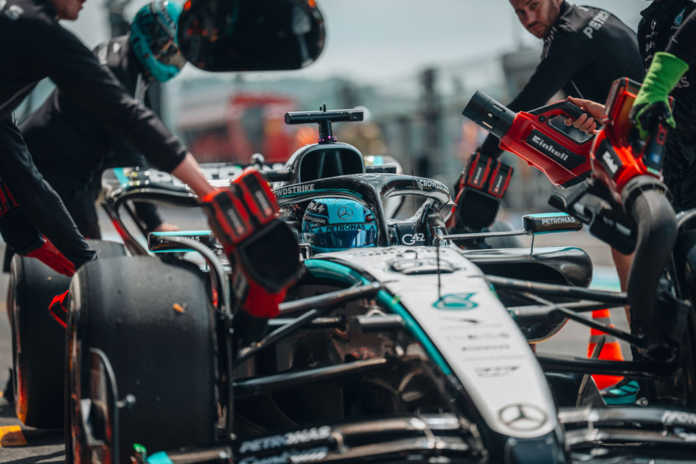En Mercedes-AMG PETRONAS Formula One-bil under et pitstopp. Teammedlemmer bruker trådløse blåsemaskiner fra Einhell for å kjøle ned bilens komponenter. Teamet er fokusert og jobber presist.
