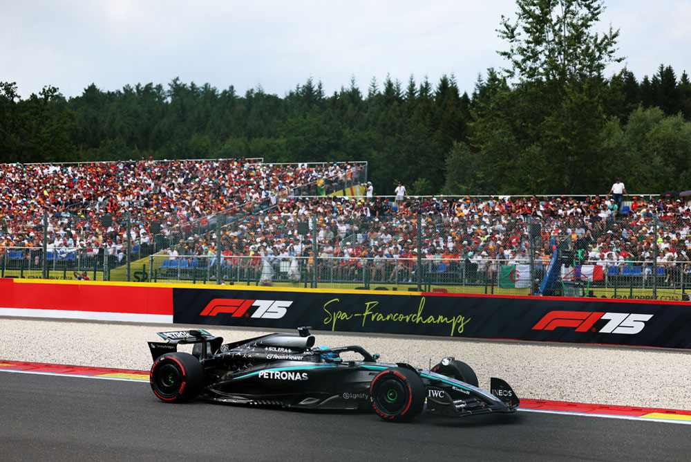 En Mercedes-AMG PETRONAS Formula One-bil kjører på Spa-Francorchamps-banen. I bakgrunnen er tribuner fulle av fans som veiver med internasjonale flagg. Spa-Francorchamps-logoen er synlig på barrieren.