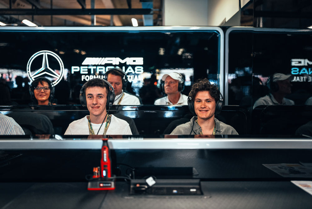 To menn sitter i Mercedes-AMG PETRONAS Formula One Teams garasje, med hodetelefoner på, og nyter opplevelsen bak kulissene. En rød Einhell-enhet er synlig i forgrunnen.