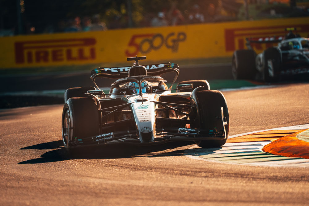 En Mercedes-AMG PETRONAS Formula One-bil kjører på en solfylt bane under et Grand Prix. Bilen tar en sving i høy hastighet med en annen bil i bakgrunnen.