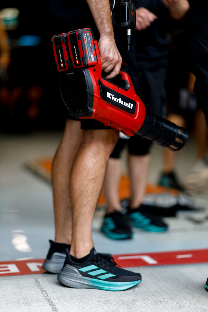Et teammedlem holder en rød trådløs blåsemaskin fra Einhell med to Power X-Change-batterier. Scenen finner sted i pit-området under et Formel 1-arrangement.