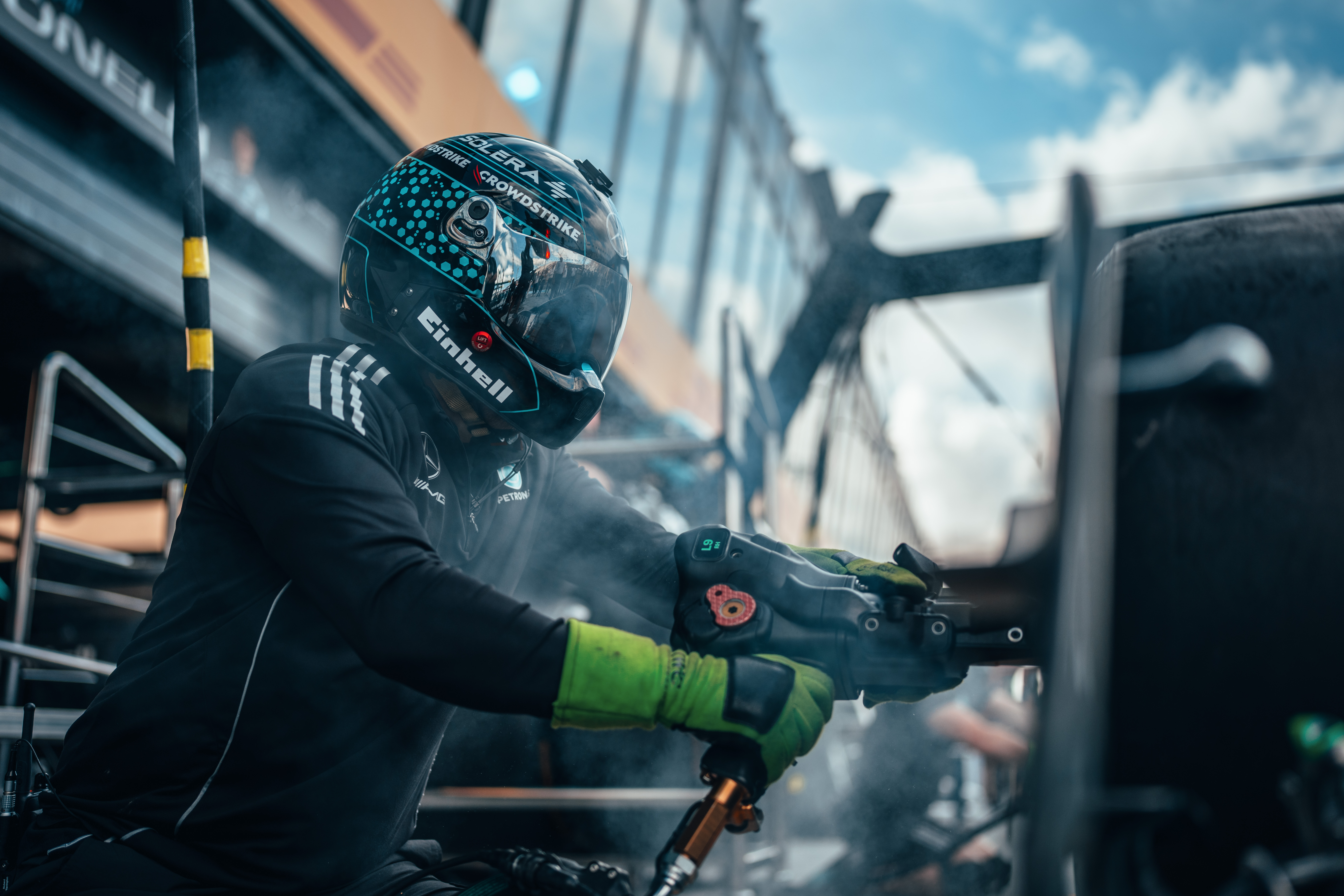 Et medlem av pitcrewet i Formel 1, iført hjelm og hansker, bruker en pneumatisk hjulmuttertrekker under et pitstopp, omgitt av røyk og utstyr.