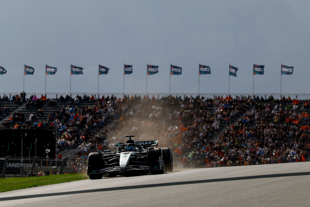 En Mercedes-AMG PETRONAS Formula One-bil kjører forbi fulle tribuner under et løp. Fans veiver med flagg i bakgrunnen og skaper en livlig atmosfære.